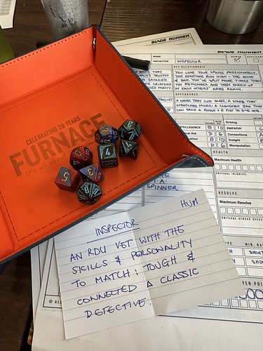 A photo of a Blade Runner character sheet with an index card saying INSPECTOR - AN RDU VET WITH THE SKILLS & PERSONALITY TO MATCH: TOUGH & CONNECTED. A CLASSIC DETECTIVE. An orange Furnace Dice Tray is adjacent with the polyhedral dice needed for Blade Runner.
