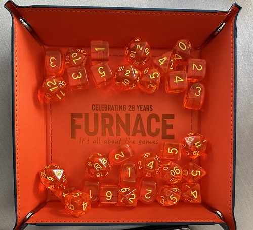 A bright orange square dice tray full of orange translucent polyhedral dice with yellow number inscribed on them. The Furnace XX logo is laser etched into the tray in the centre of the base. It says Celebrating 20 Years - Furnace - It's all about the Games.